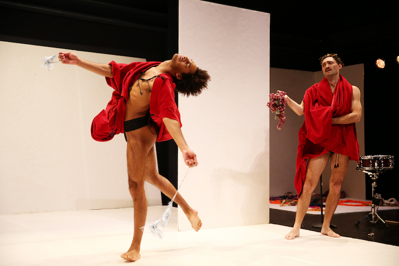 Two men wearing berry-hued robes and black underwear. One tosses his head back as he stands on one leg. While the other stands watching holding grapes in one hand.